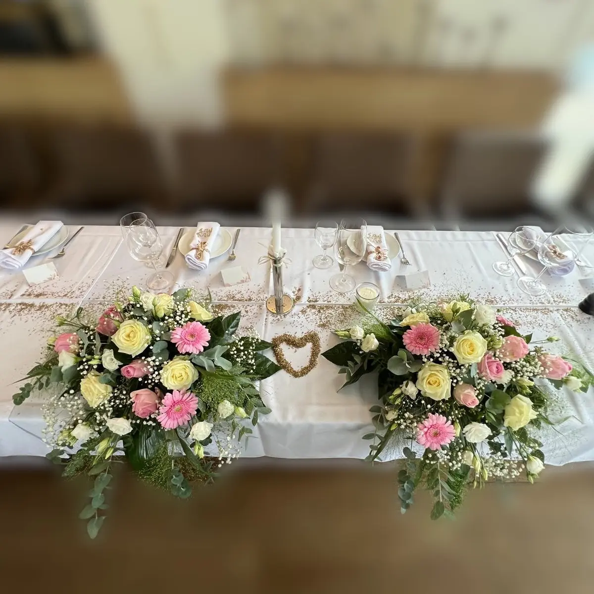 Festtafel mit zwei Gestecken aus Rosen, Gerbera, Eukalyptus und Schleierkraut, dazwischen ein Herz