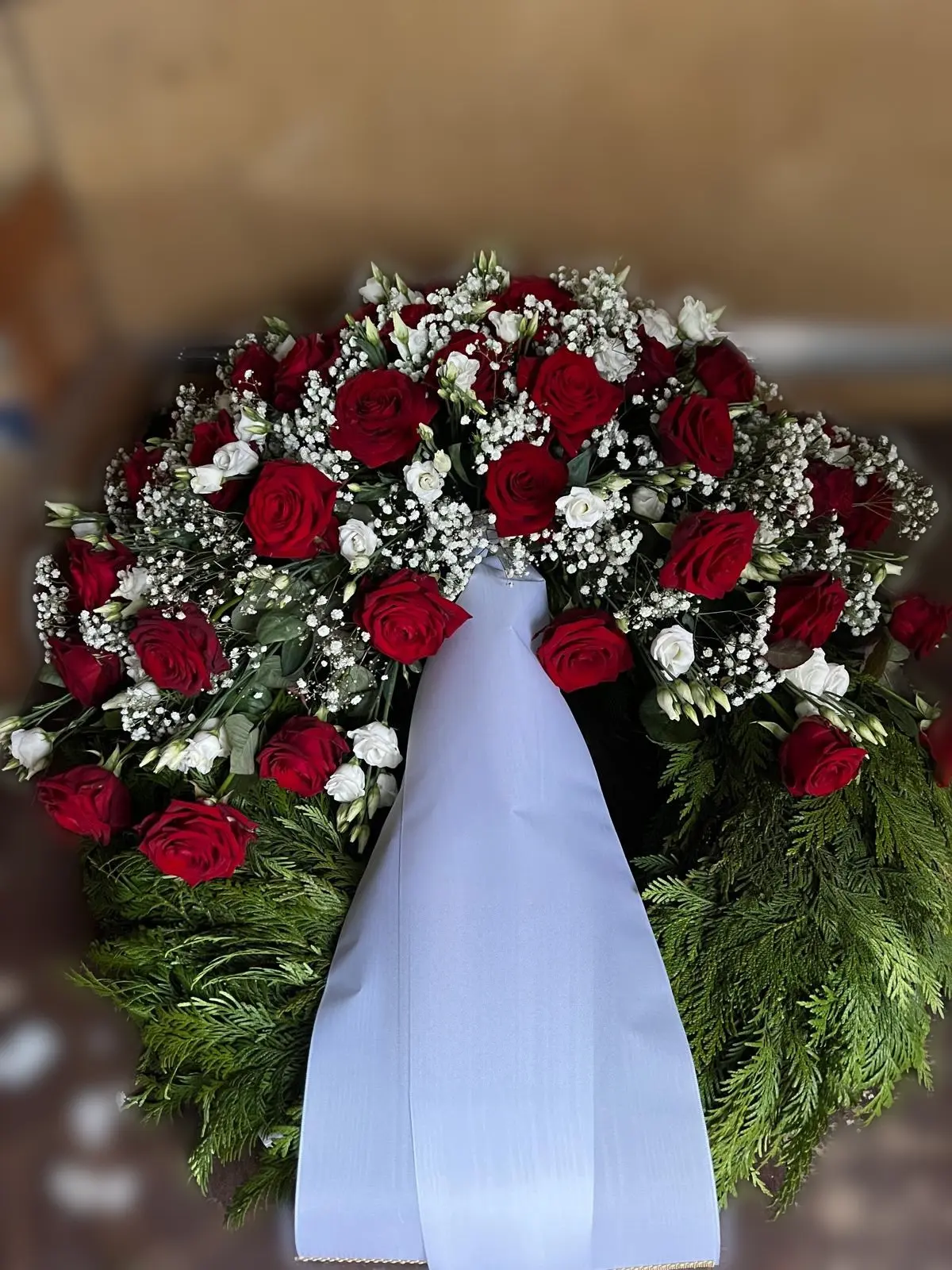 Trauergesteck aus roten Rosen, weißem Lisianthus und Schleierkraut auf Koniferengrün, mit weißer Schleife